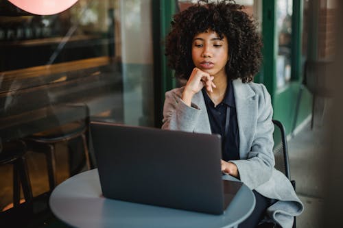 Concentrated African American female sitting at table in outdoor cafeteria and leaning on hand while browsing computer during online work