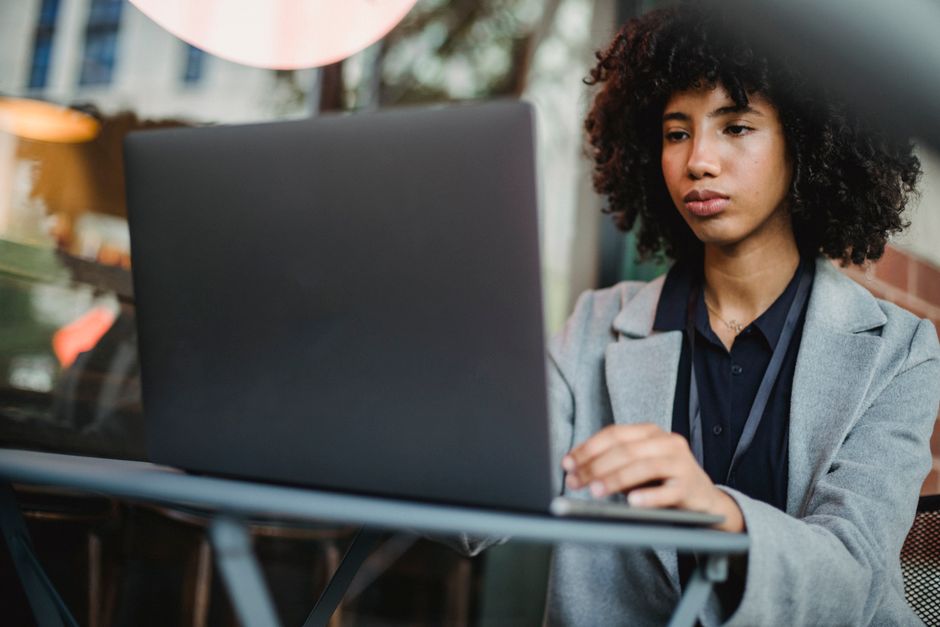 Serious African American female freelancer in elegant suit working on computer while sitting at table in outdoor cafe with blurred background