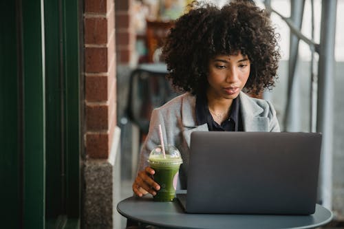 Attentive black businesswoman watching laptop with beverage in cafe