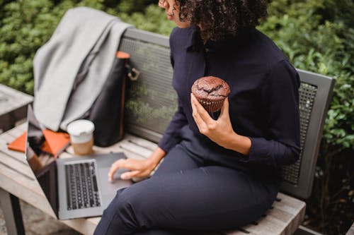 High angle of crop ethnic female distance worker browsing internet on netbook while sitting on street bench with tasty muffin