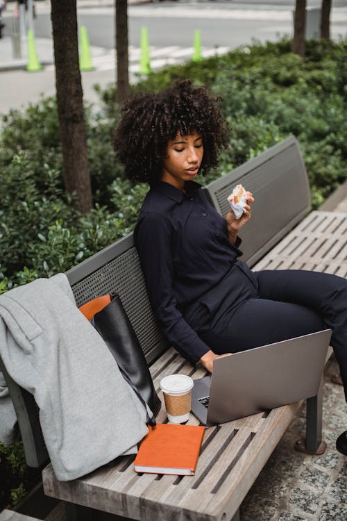 From above side view of young black female distance worker browsing internet on netbook while resting on urban bench with puff