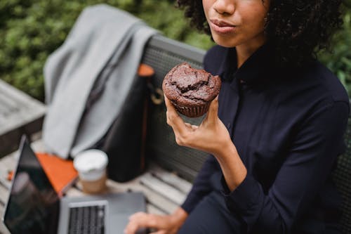 Crop black remote employee with tasty muffin and laptop outdoors