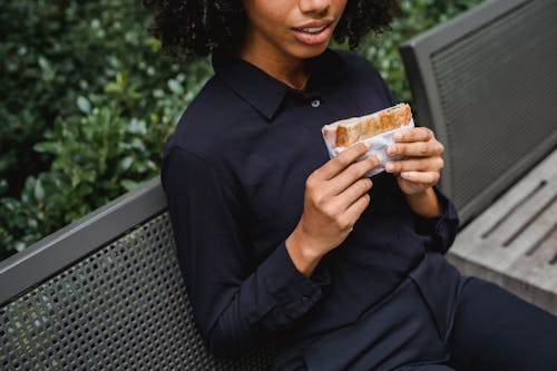 Free Crop faceless African American female in stylish clothes sitting on bench in park while holding sandwich Stock Photo
