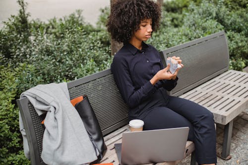 Crop African American woman in trendy outfit sitting on bench with netbook and coffee while eating delicious snack in park
