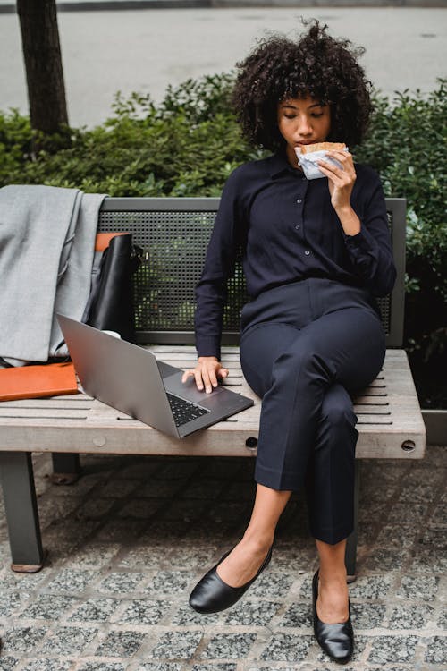 Busy African American woman in formal clothes sitting on bench and browsing netbook while having lunch in park