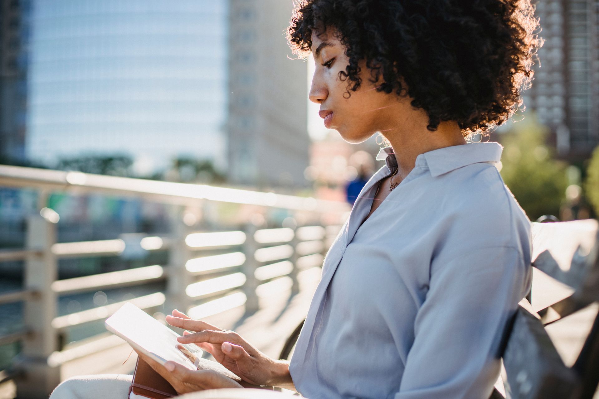 A woman relaxes on a bench outdoors in the city, using a digital tablet under bright daylight.