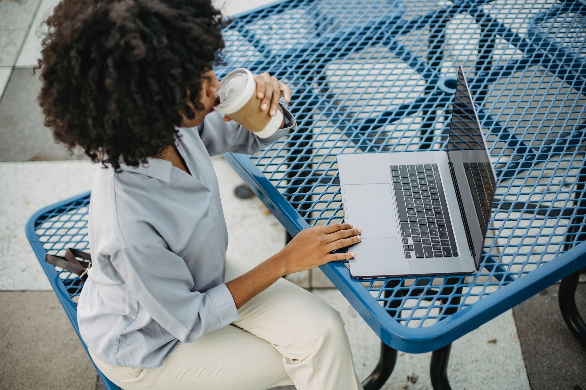 Woman with curly hair drinking coffee and working on a laptop at an outdoor table.