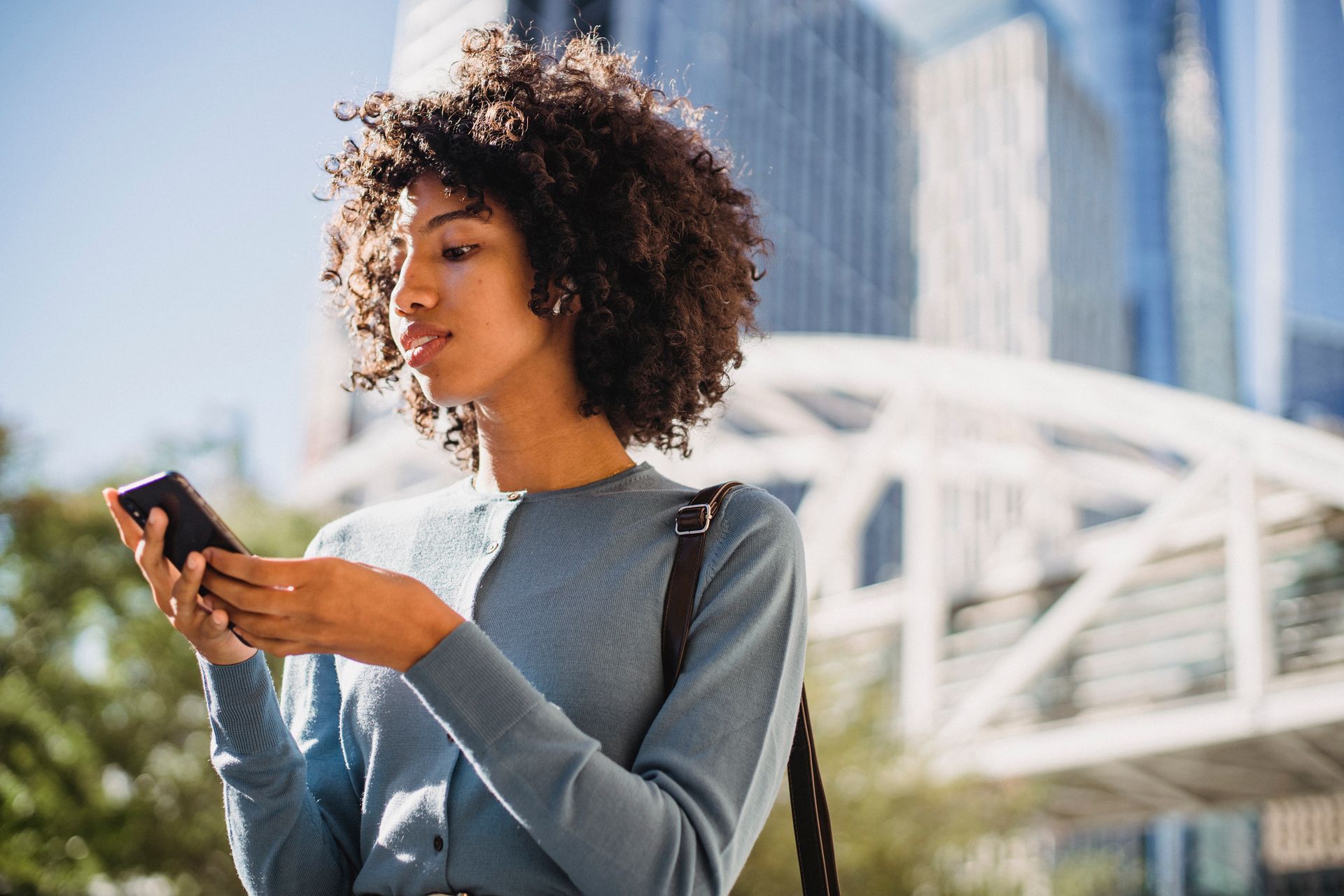 Stylish young woman with curly hair using a smartphone outdoors in a city setting.