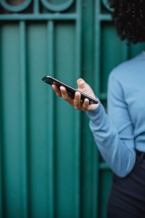 Free A person in a blue sweater holds a smartphone against a teal urban backdrop. Modern lifestyle concept. Stock Photo