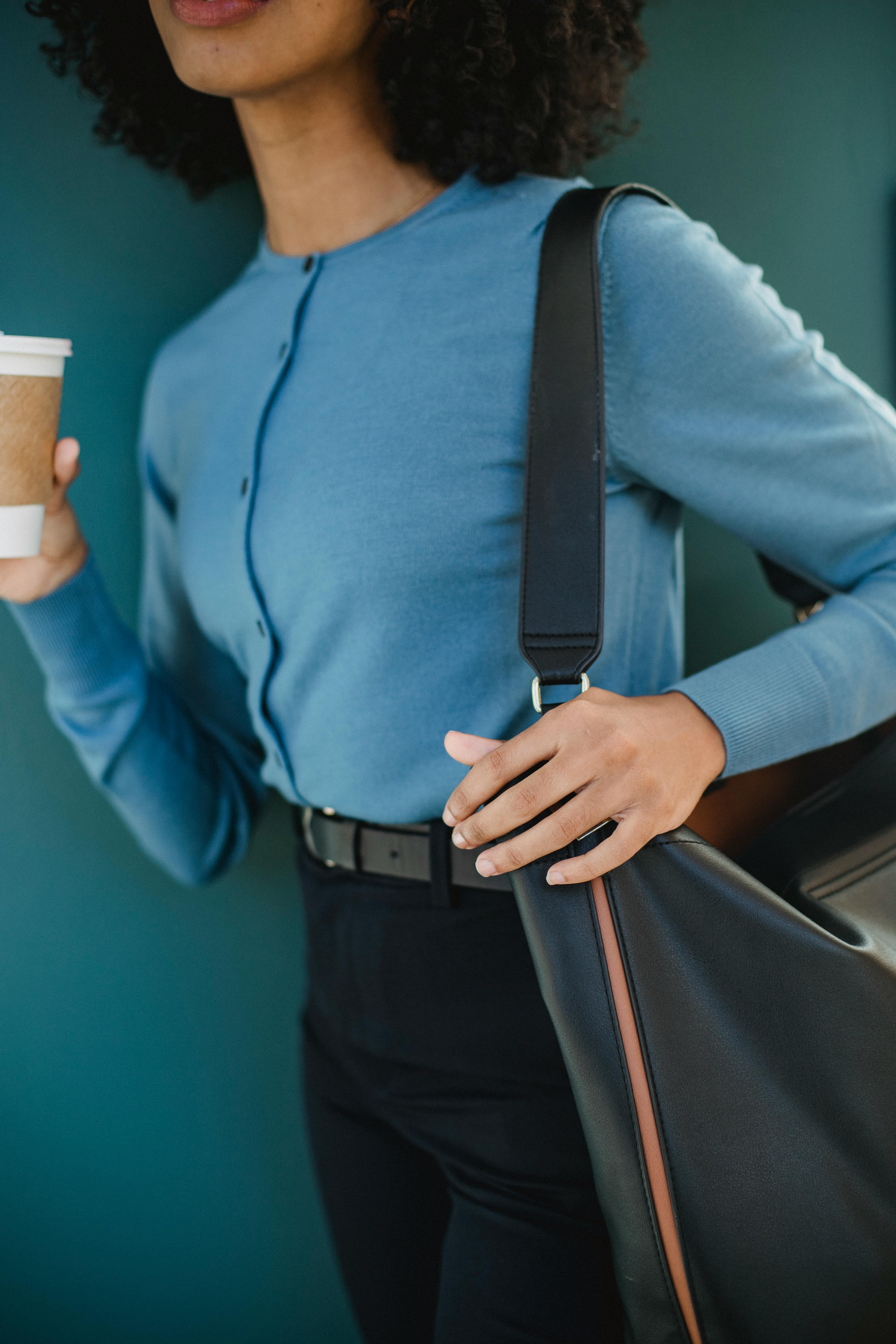 Free Fashionable woman in blue sweater holding a coffee cup while carrying a bag, showcasing modern urban style. Stock Photo