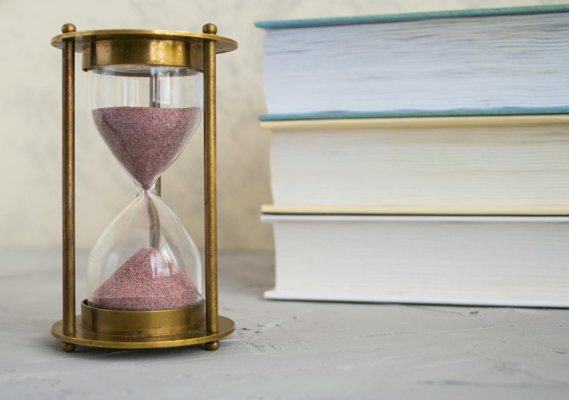 Close-up of hourglass with red sand beside stacked books, symbolizing time and knowledge.