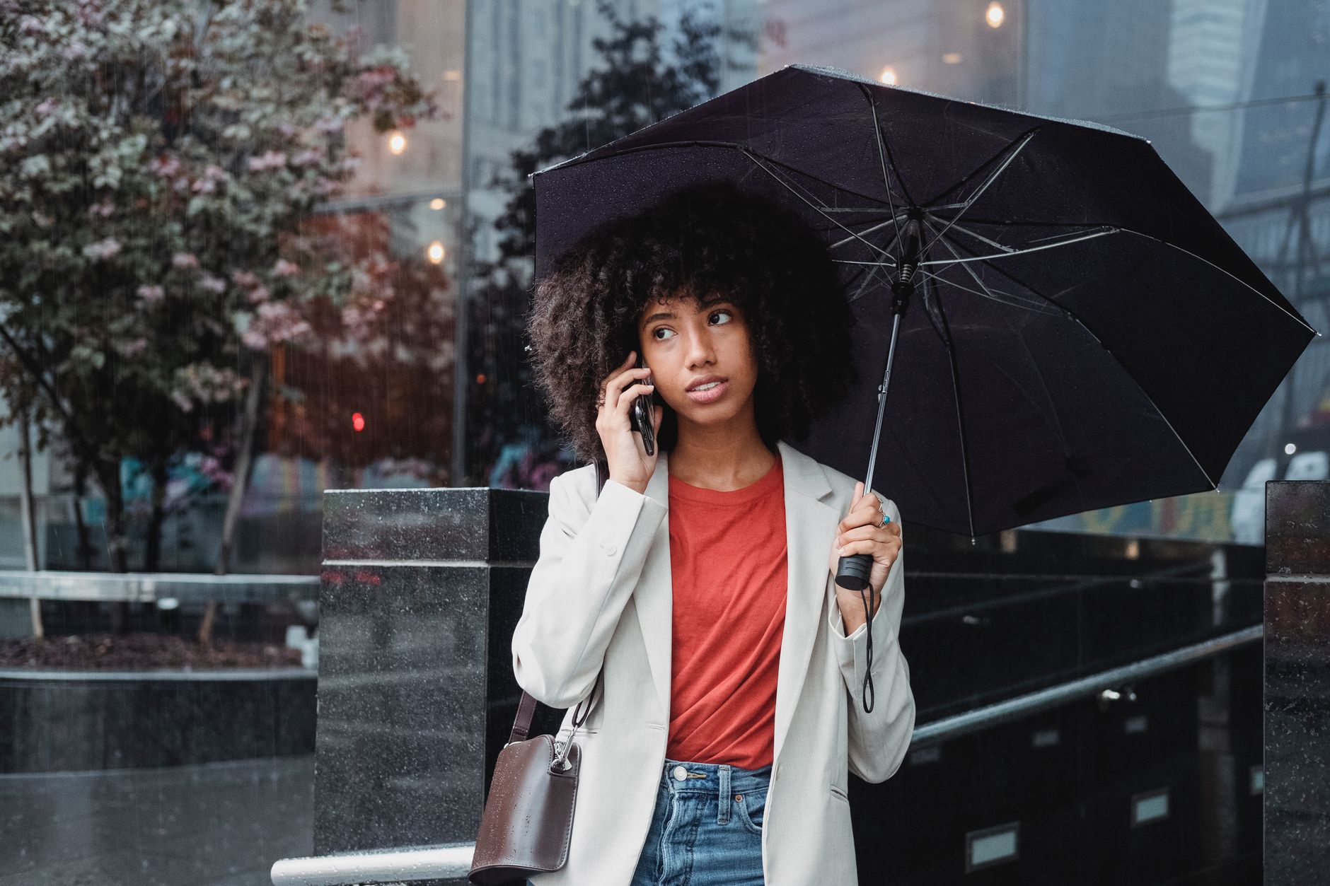 Stylish young woman with curly hair using smartphone under umbrella in urban setting.