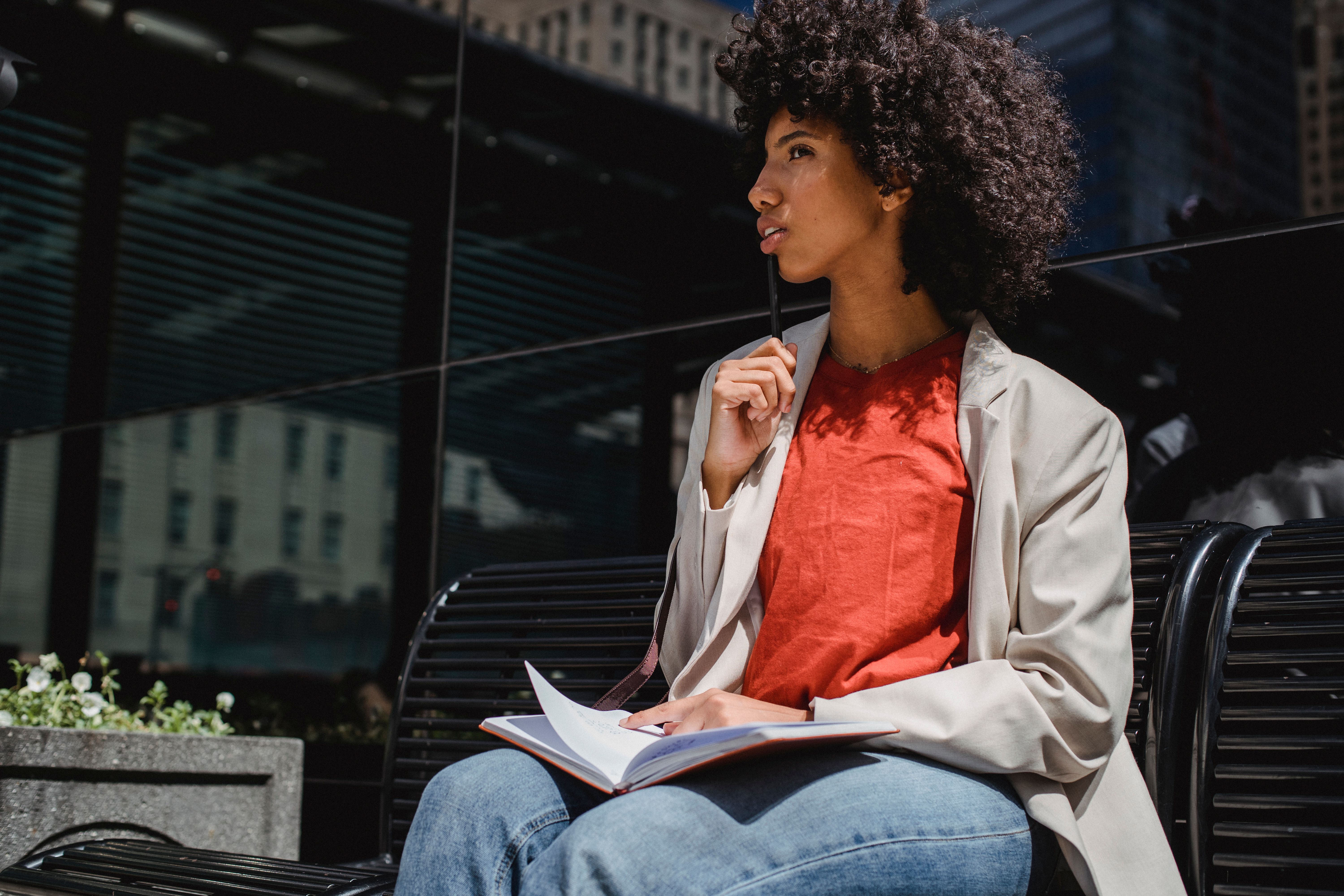 Woman sits outdoors on a bench, writing in a notebook in a city environment.