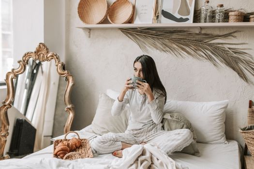 Woman in pajamas enjoys coffee and pastries in a stylish and cozy bedroom setting.
