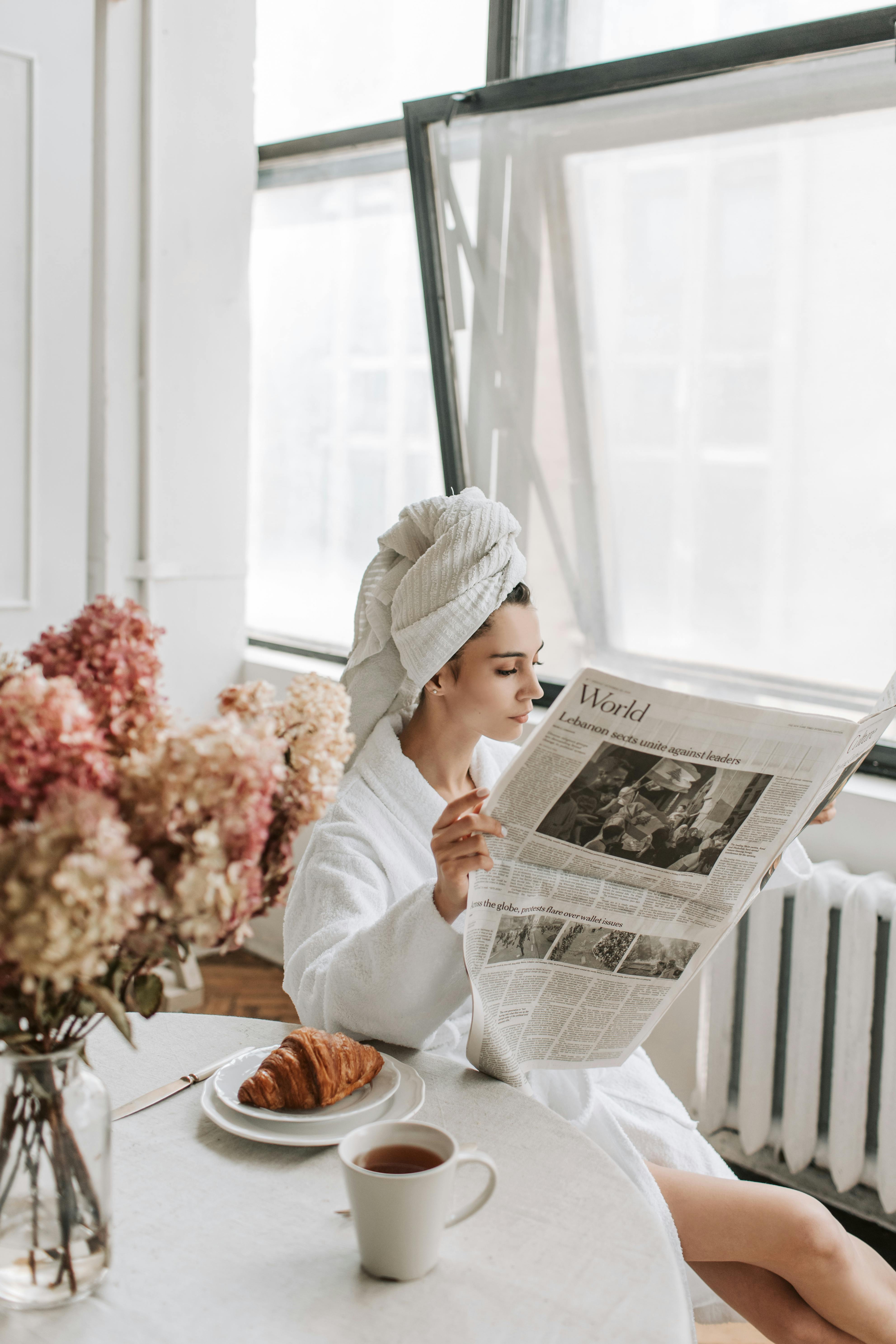 woman in bathrobe reading a newspaper while sitting at the table