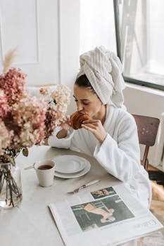 Relaxed morning scene of a woman in a bathrobe eating a croissant and drinking coffee indoors.