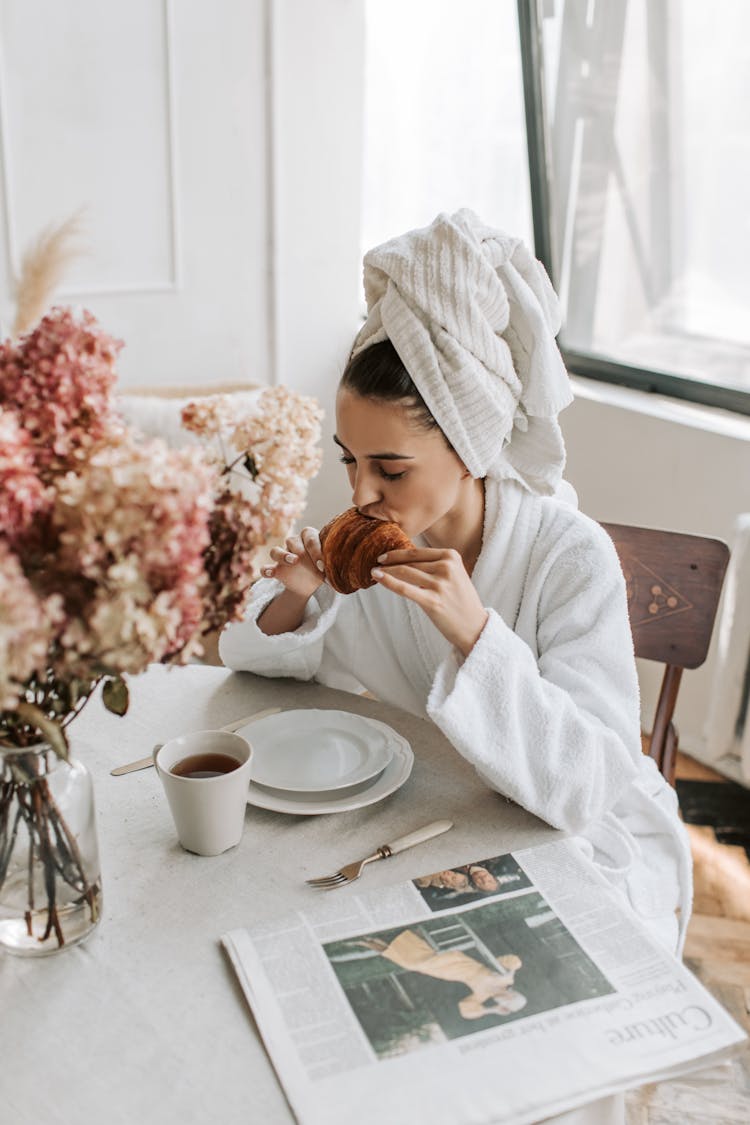 A Woman In Bathrobe Eating Croissant