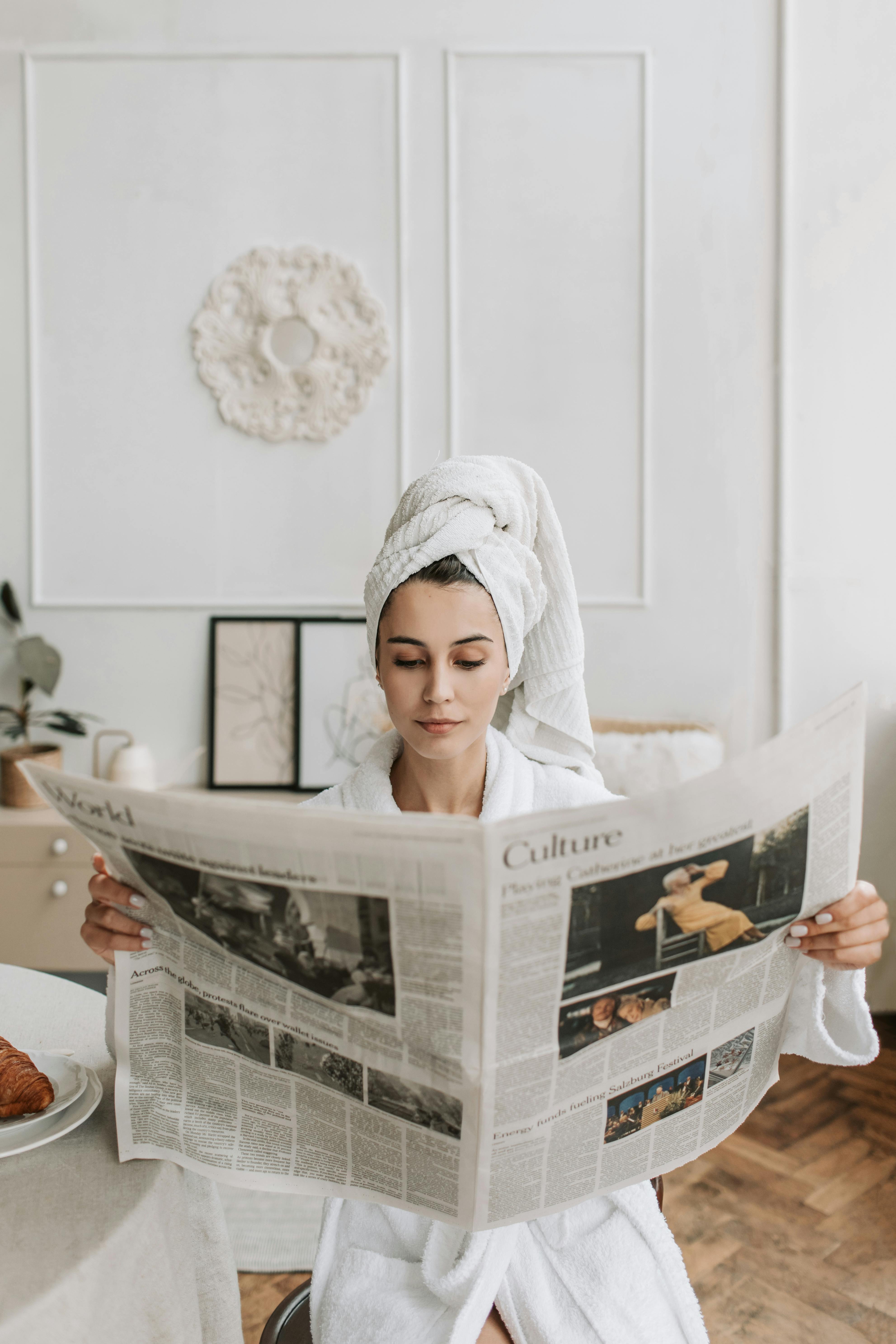 woman in white robe and head towel seriously reading the newspaper