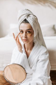 Woman in a bathrobe using eye patches in a serene indoor environment.
