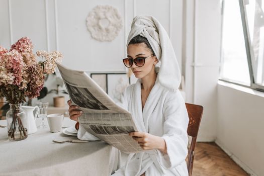 Woman in bathrobe reading a newspaper at a stylish indoor setting with flowers.