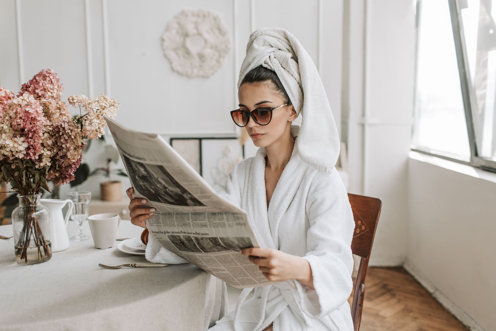 A woman in a white robe and sunglasses reading a newspaper at home — dressing for her own pleasure