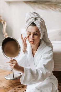 Woman in bathrobe applying skincare product using mirror in a cozy bedroom.