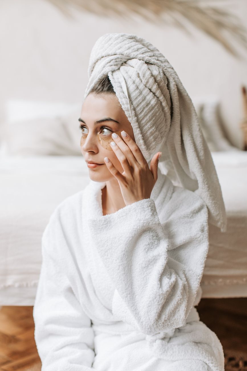 Woman in a white robe applying face cream, wrapped in a towel, sitting indoors.