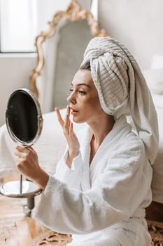 Woman in a bathrobe applying facial care in a cozy bedroom setting.