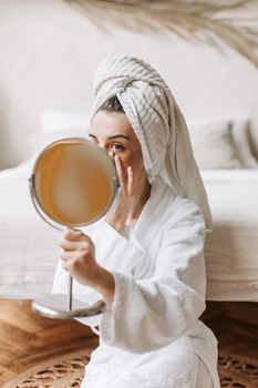 A woman in a bathrobe applies skincare while sitting in a bedroom with a towel on her head.