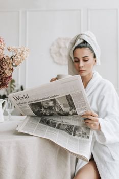 Woman in white robe reading newspaper while enjoying coffee indoors.