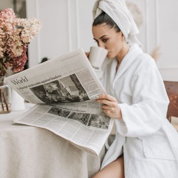 Woman in robe enjoys morning coffee while reading a newspaper indoors.