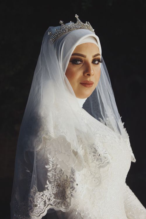 Free Side view gorgeous young Muslim bride wearing classy modest white dress and hijab covered with chic veil while looking at camera and standing on black background in studio Stock Photo