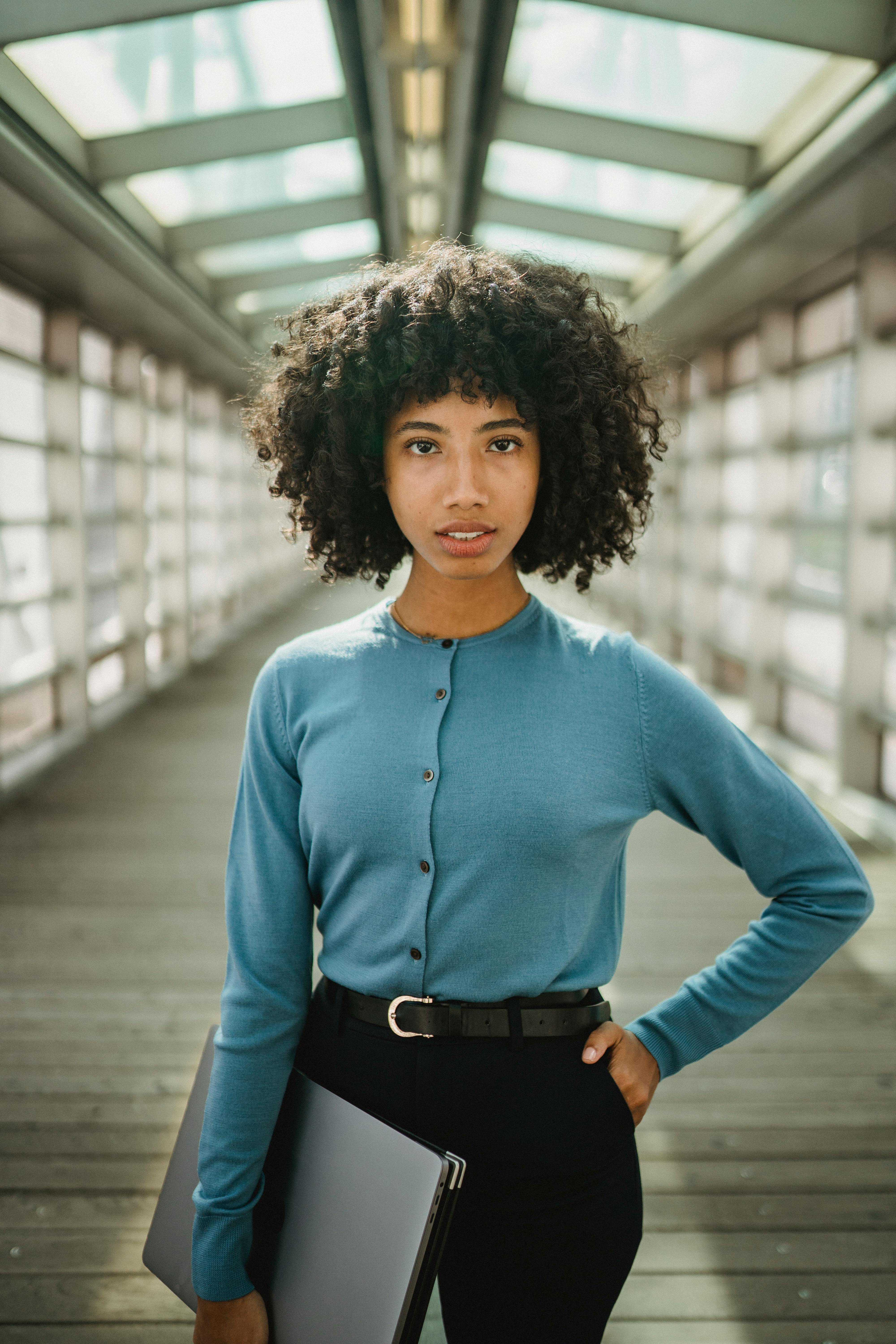 Young woman with afro hair standing confidently with a laptop in a modern corridor.