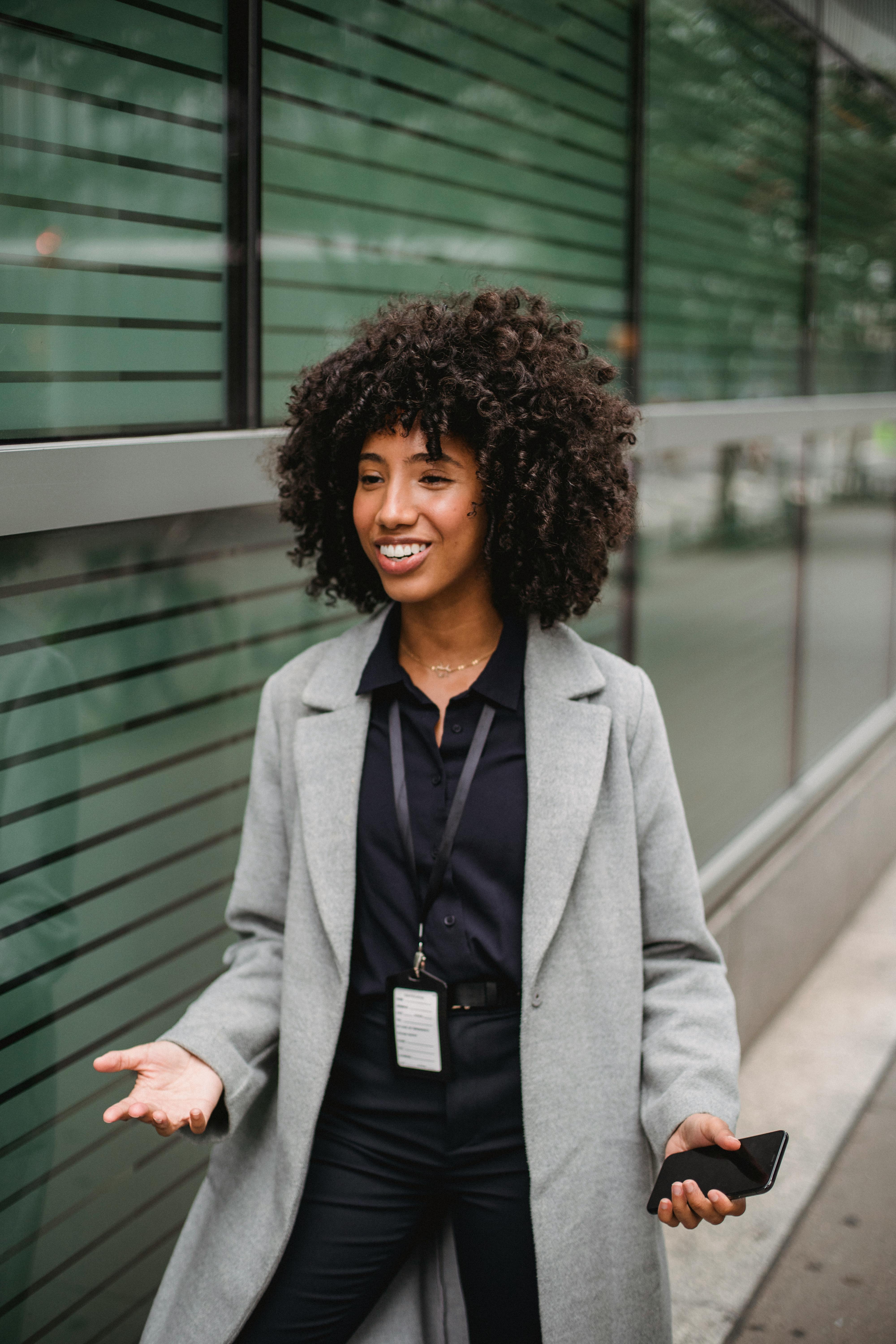 Businesswoman in a gray coat standing outside a glass building, holding a mobile phone and smiling.
