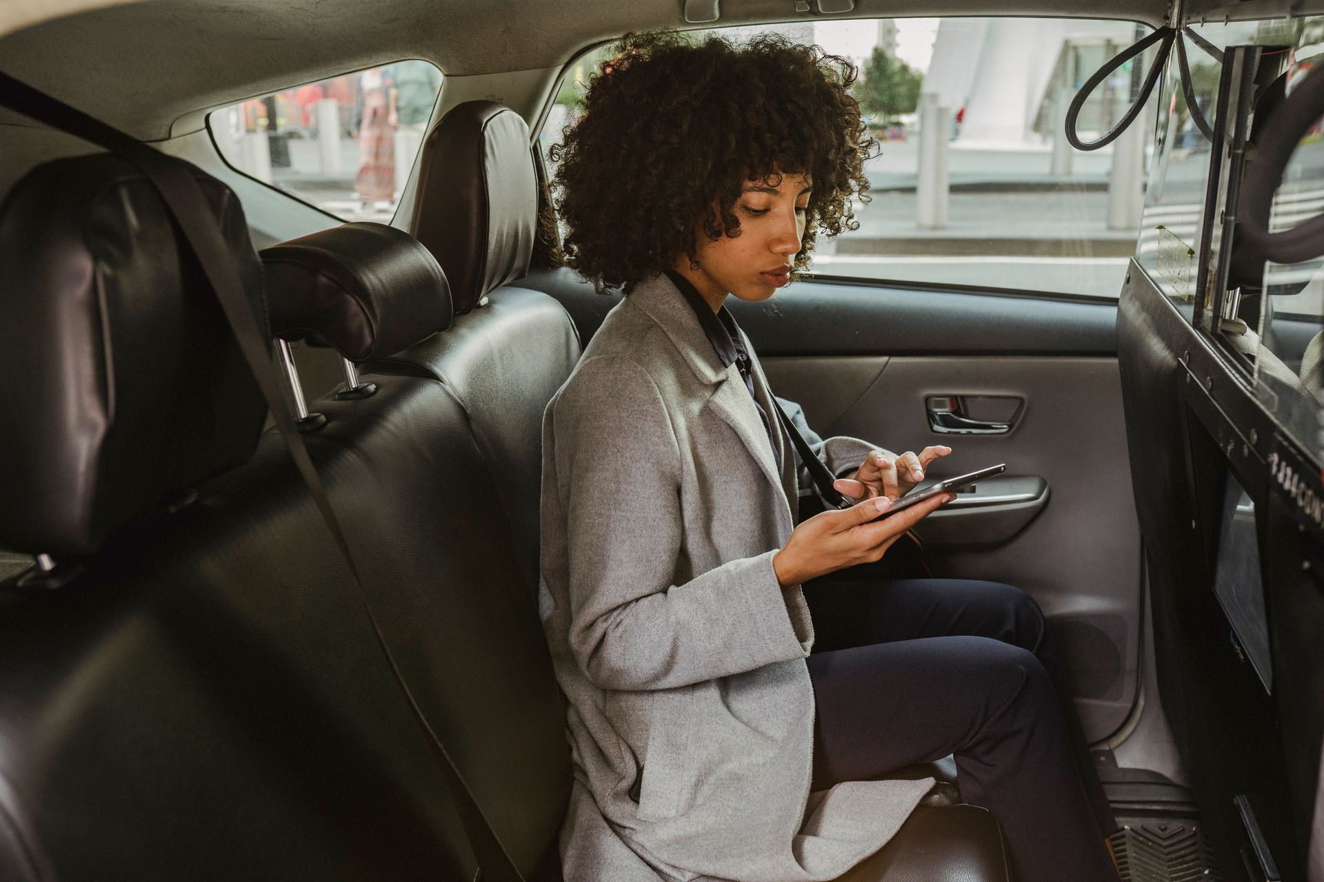 Woman with curly hair using a smartphone inside a car with black leather seats.