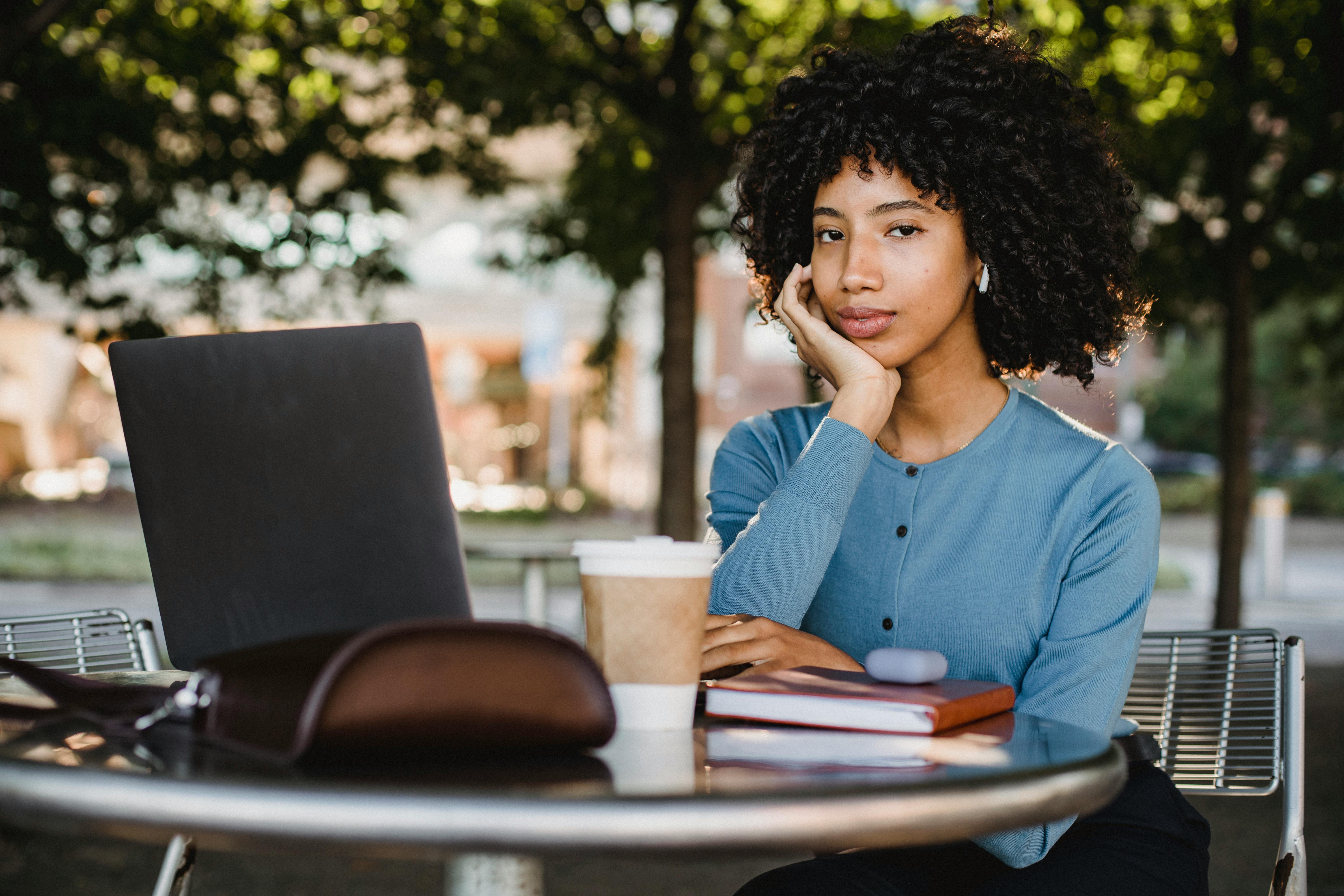 African American woman with curly hair working on a laptop at an outdoor cafe table.