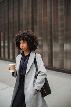 Confident woman walking on street holding coffee, wearing a gray coat and afro hairstyle.