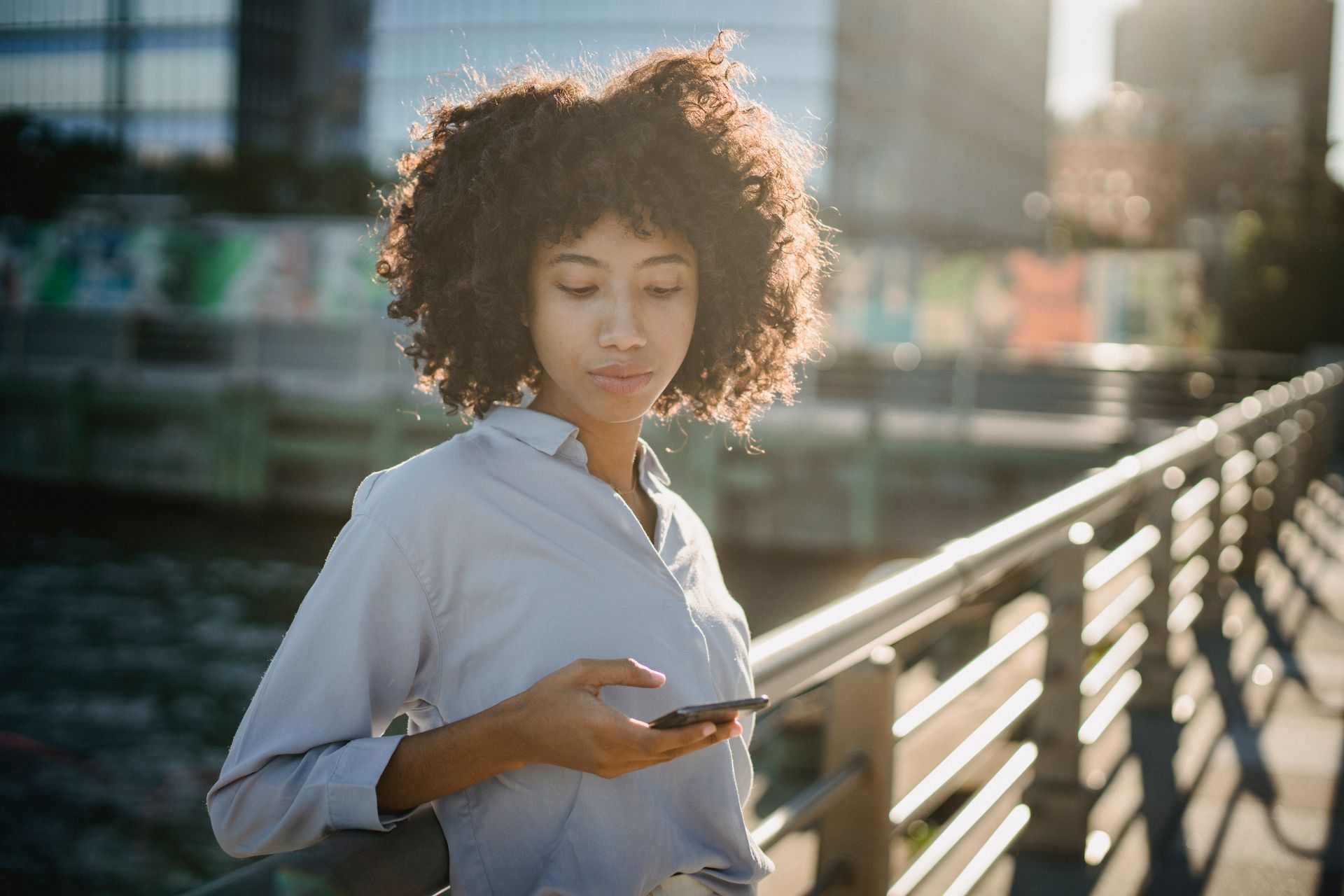 Portrait of a young woman with curly hair using her smartphone on an urban bridge during daylight.