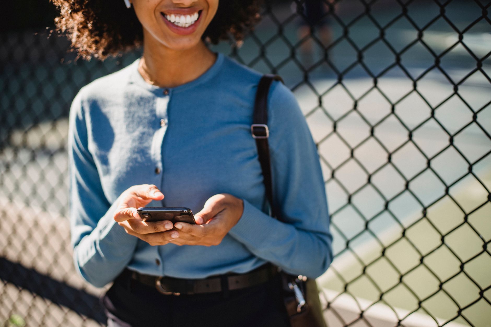 A cheerful woman uses her smartphone outdoors near a fence.