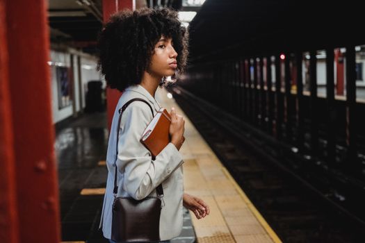 African American woman with afro hair holding a notebook while waiting at a subway station.