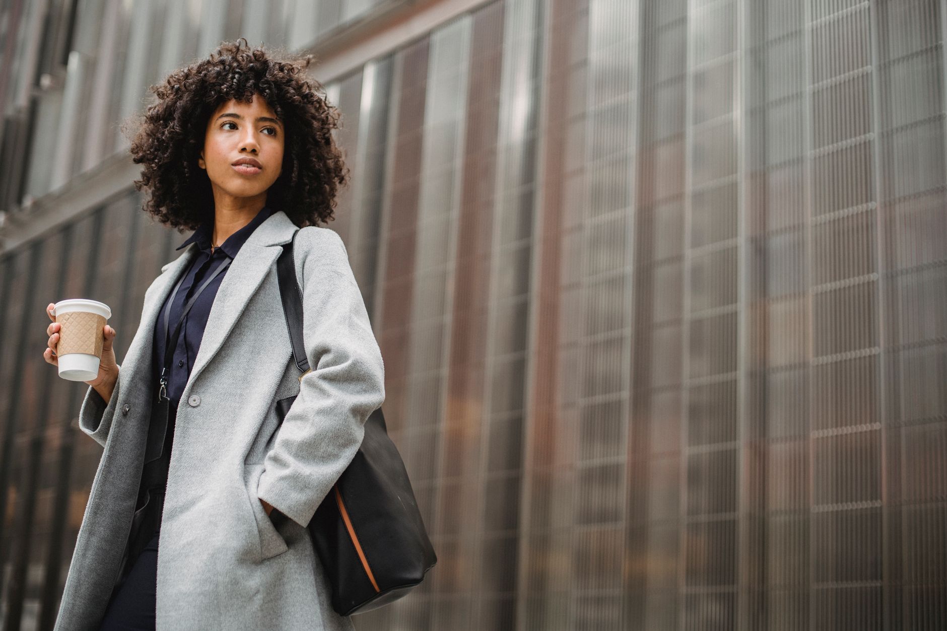 A confident woman in a gray coat holding coffee on a city street, walking with easy composure