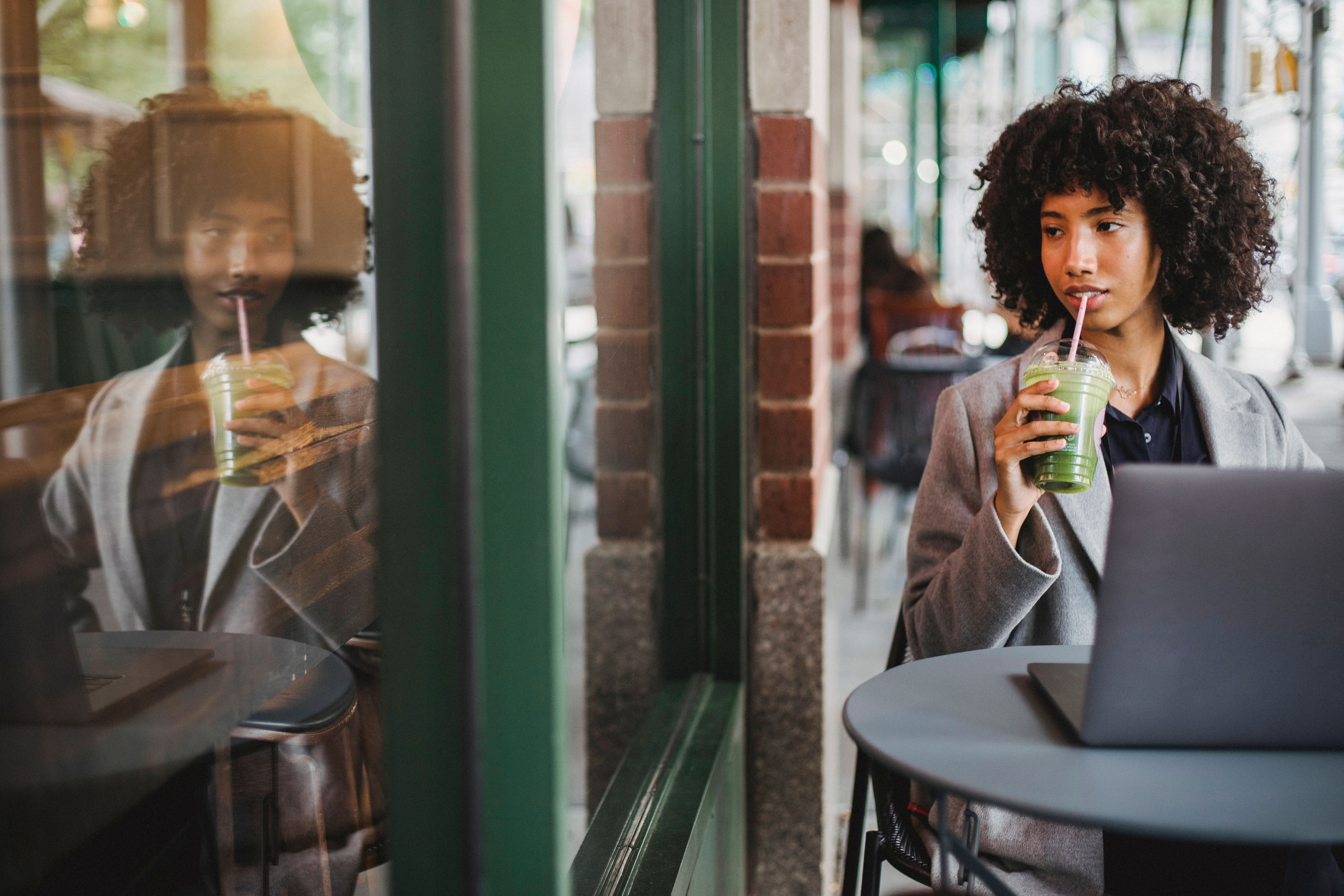 Elegant woman sipping green smoothie and working at a cafe table near a window.