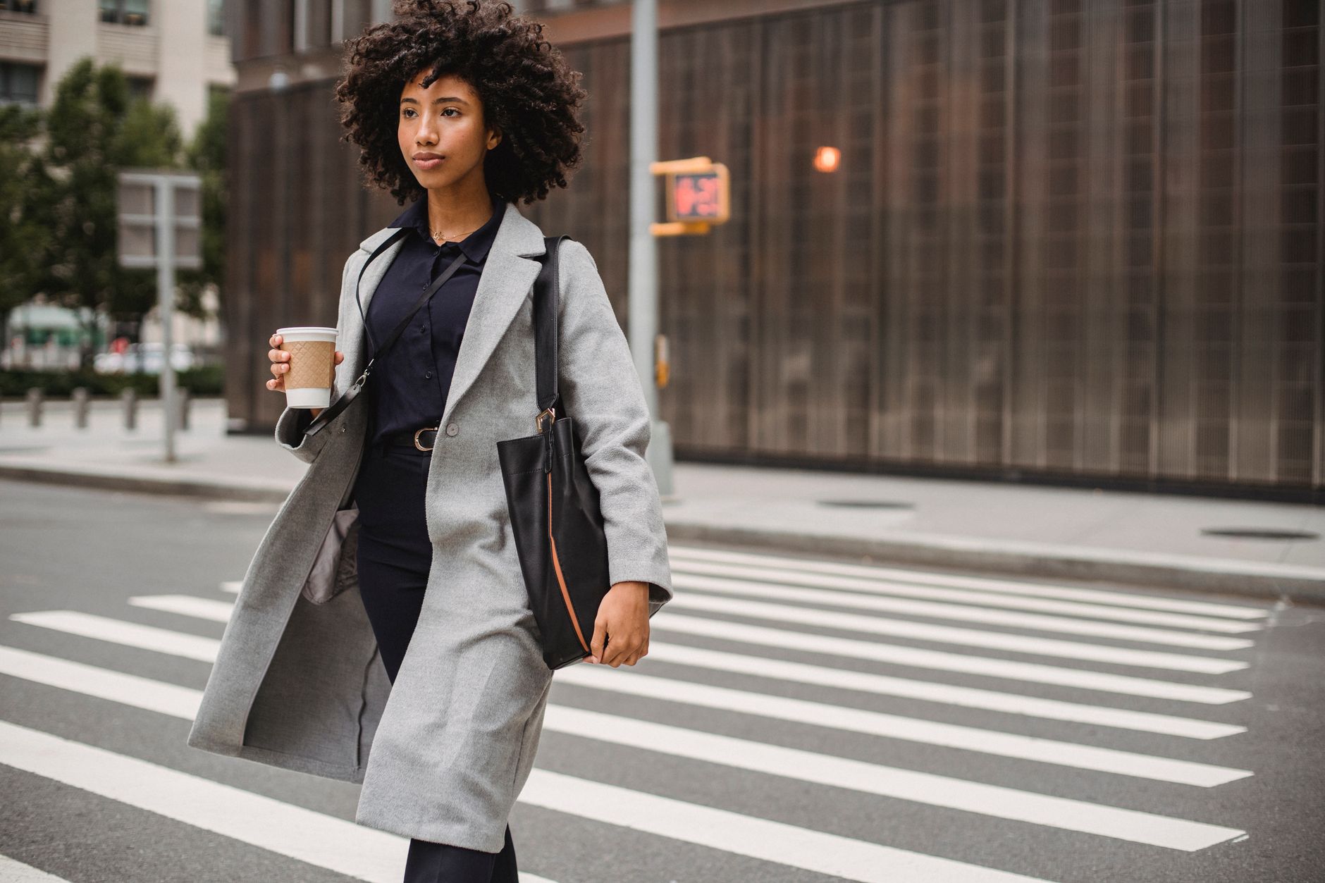 Young woman crossing an urban street holding coffee, exuding confidence in a gray coat.