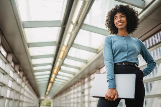 A young woman stands confidently in a bright urban hallway holding a laptop.