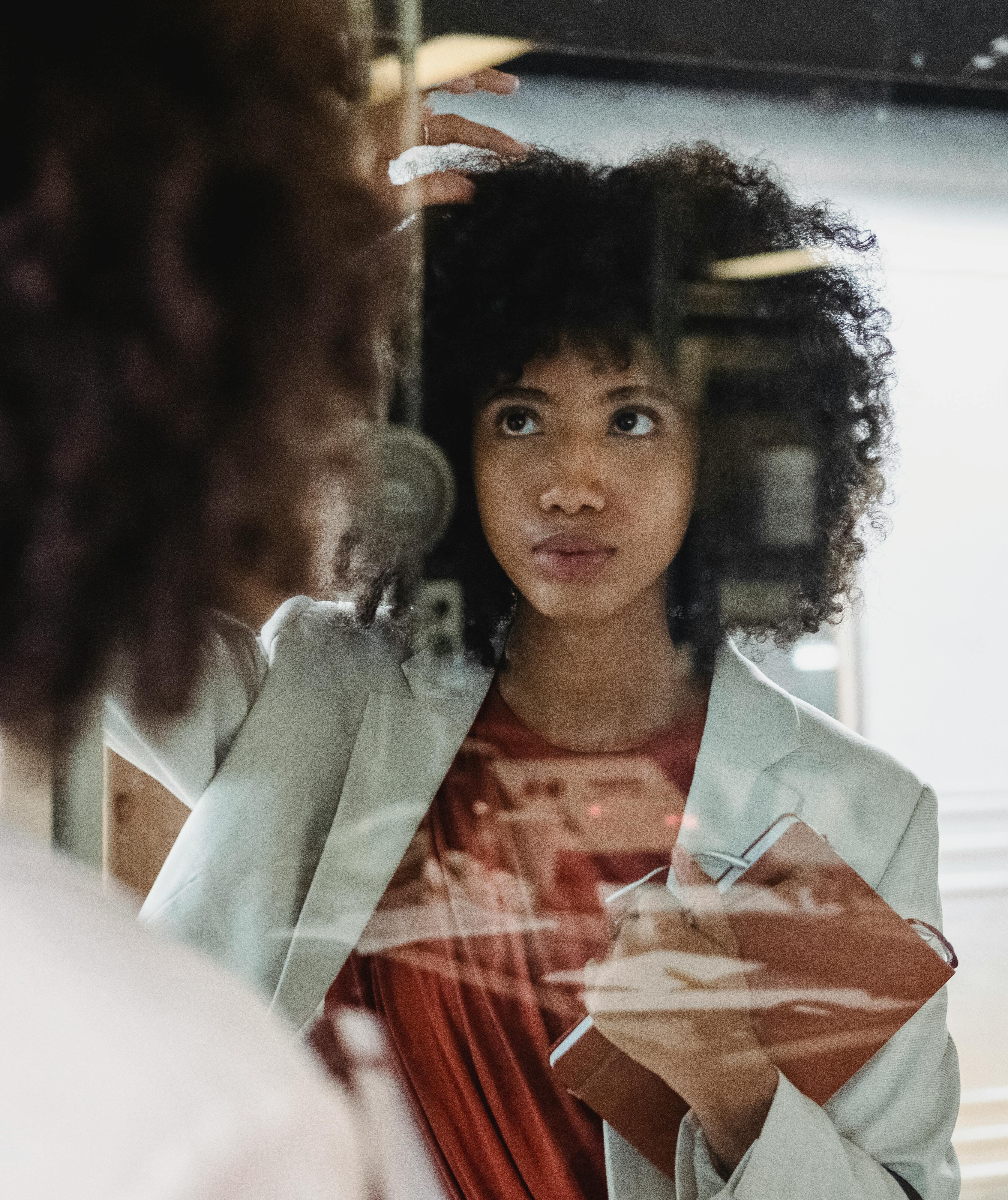 Young woman looking at her reflection, contemplating, holding a notebook indoors.