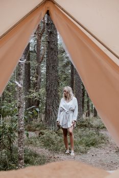 A woman in a white dress stands amid tall trees in a forest, seen from a tent.