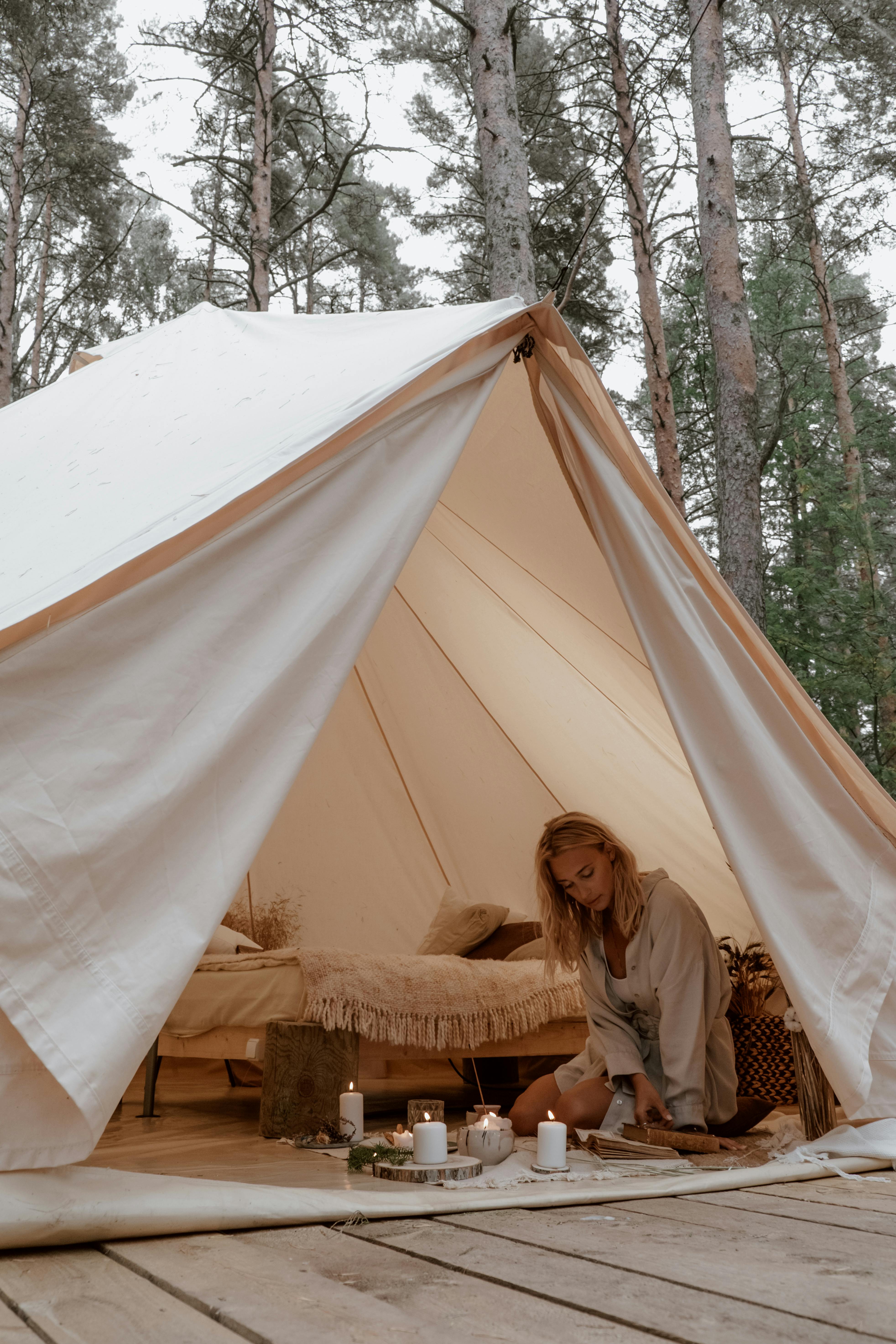 Woman kneeling in glamping tent amidst forest, enjoying candles' glow.