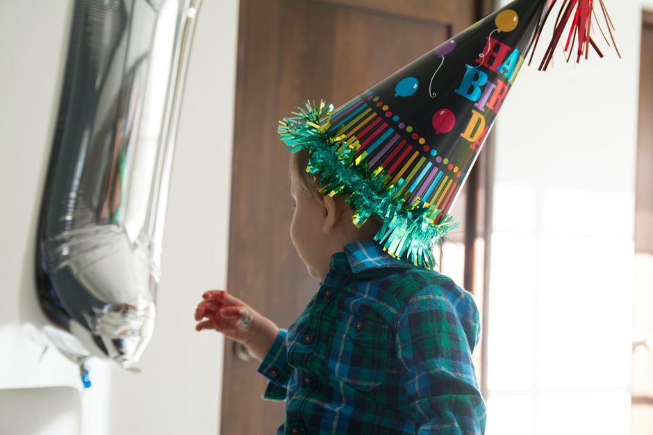 A young child wearing a colorful birthday hat indoors, next to a silver balloon.