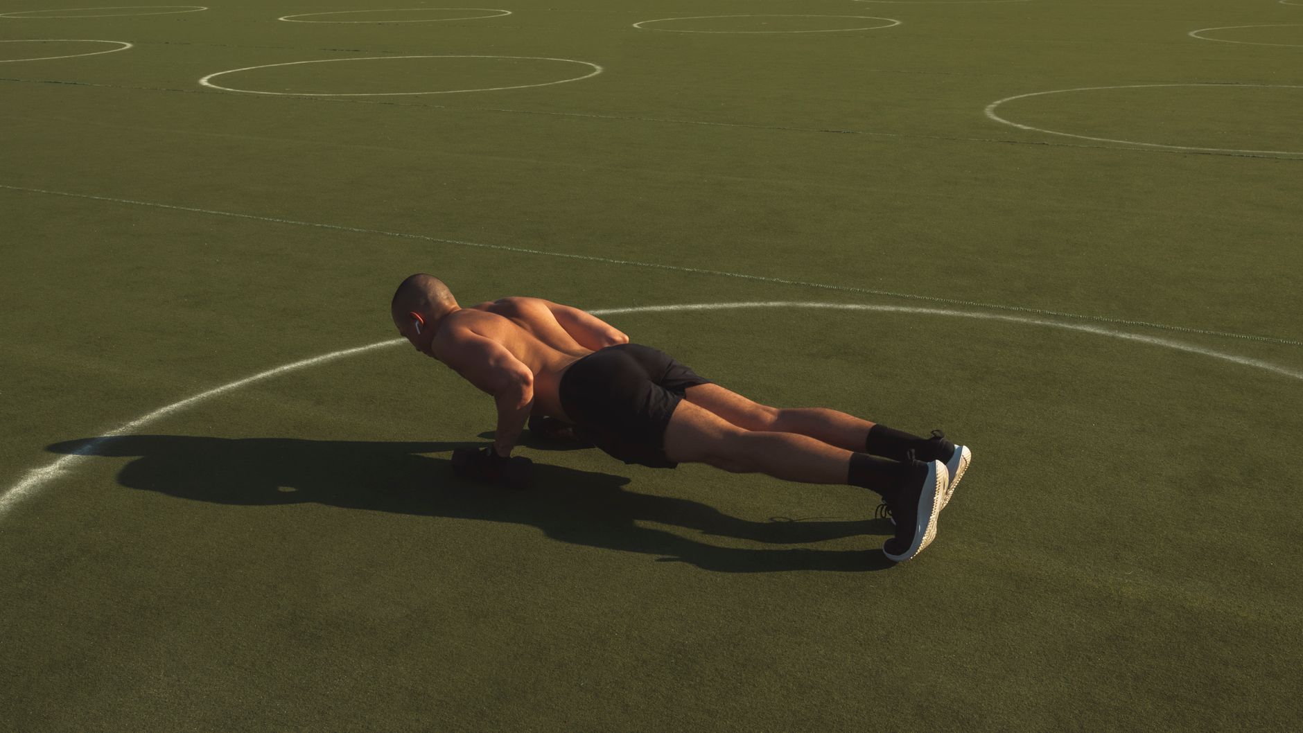 A shirtless man performing push-ups on a green field under bright daylight.
