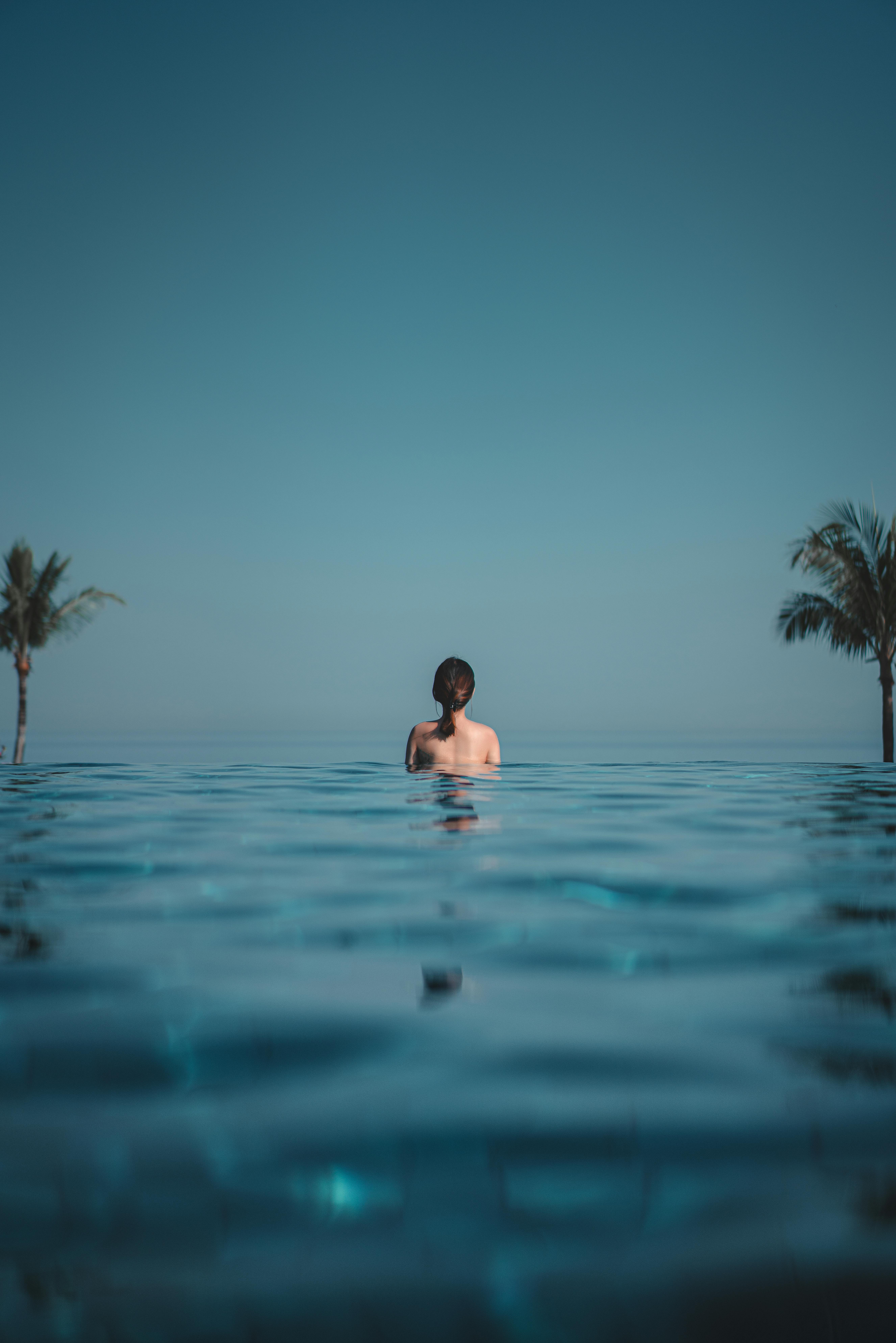 Back view anonymous slim brunette standing in blue swimming pool water in tropical resort and admiring cloudless endless blue sky